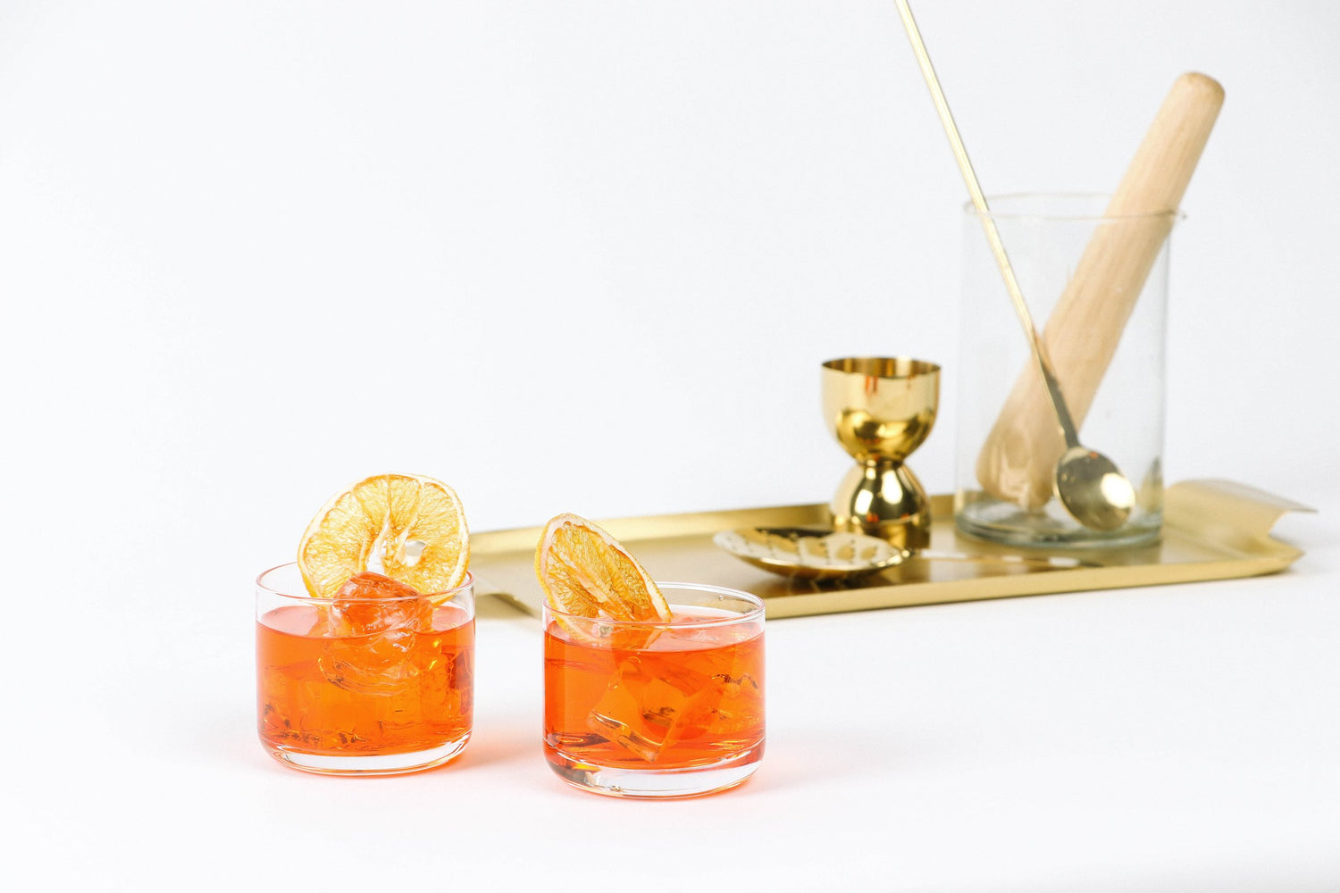 Two cups filled with an orange beverage and topped with orange sliceswith a tray in the background. Tray has cocktail-making tools.